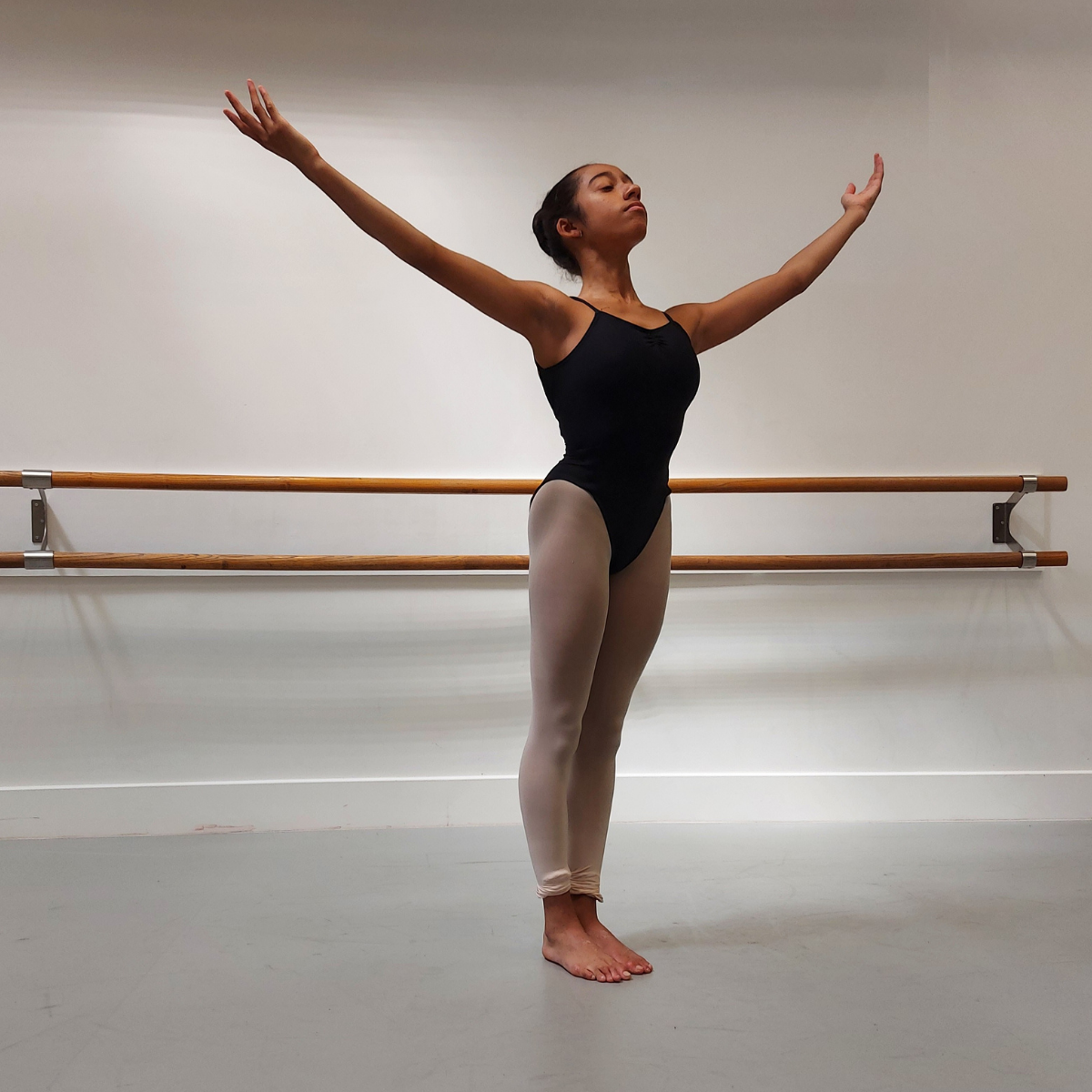 A person standing in a ballet pose wearing a black leotard with the fabric flowing around the body, including a high leg line and a front gather.